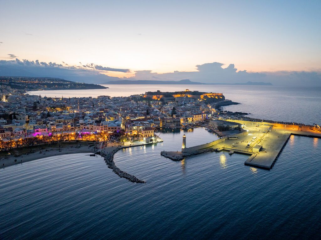 A city lights by the sea at night, with illuminated buildings reflecting in the water